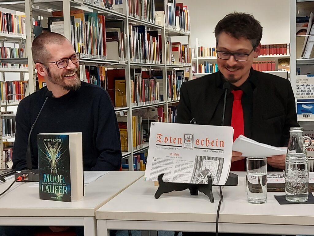 Boris Koch und Carsten Schmitt sitzen  nach ihrer gemeinsamen Lesung lachend vor Bücherregalen
